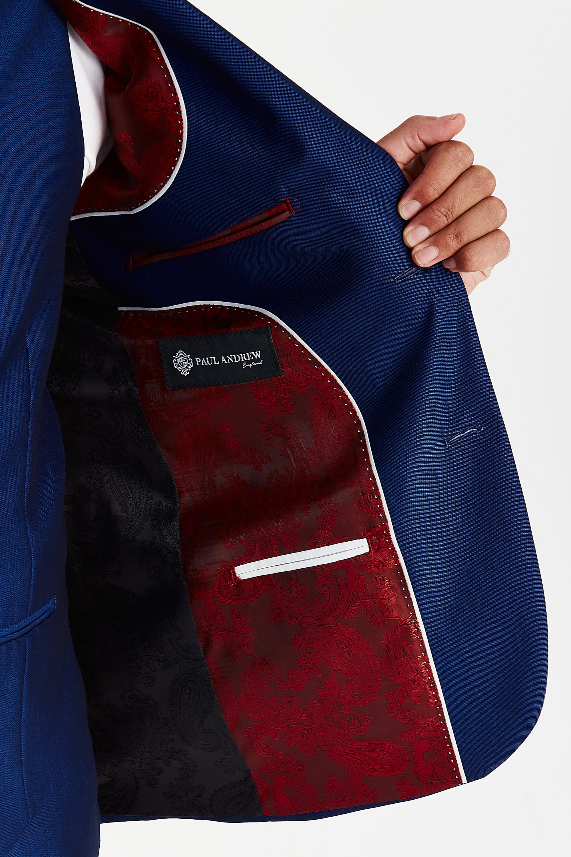 Detail view of a blue suit jacket interior with red and black paisley lining, white piping, inner pockets and a Paul Andrew label.