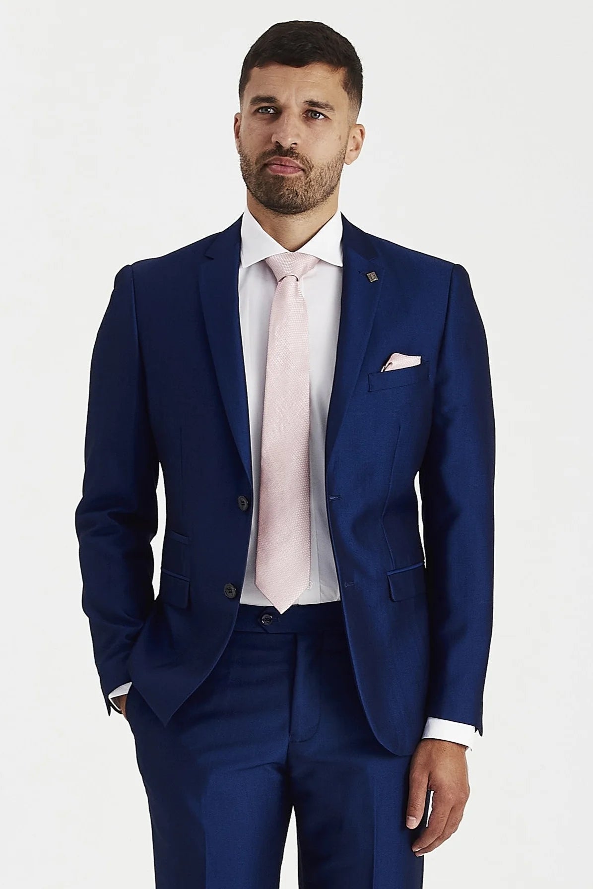 Knee-up view of a man wearing a glossy blue suit blazer and trousers, styled with a white shirt, a light pink tie, and a matching pocket square.