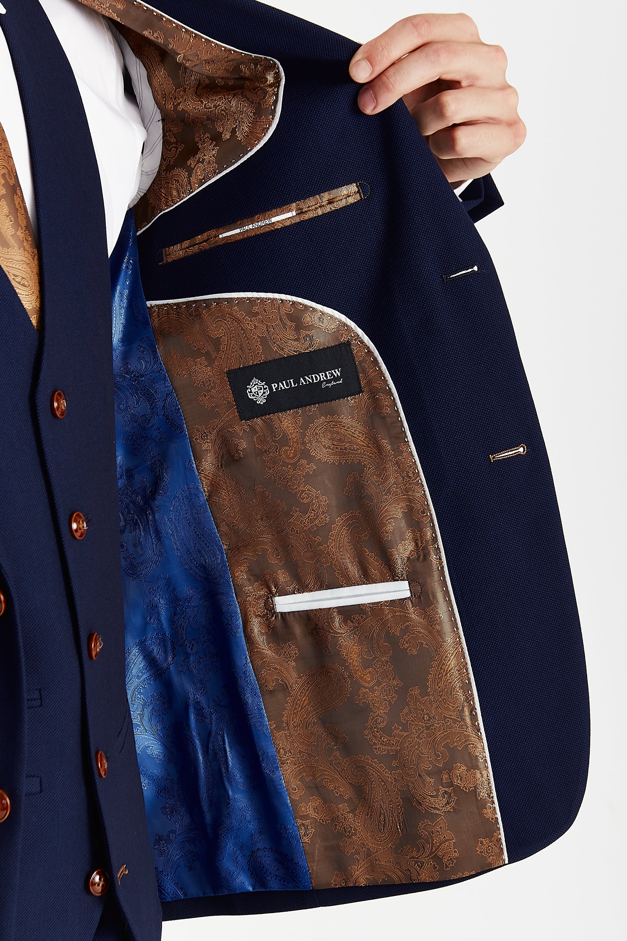 Detail view of a navy textured suit jacket interior with brown and blue paisley lining, white piping, inner pockets and a Paul Andrew label.