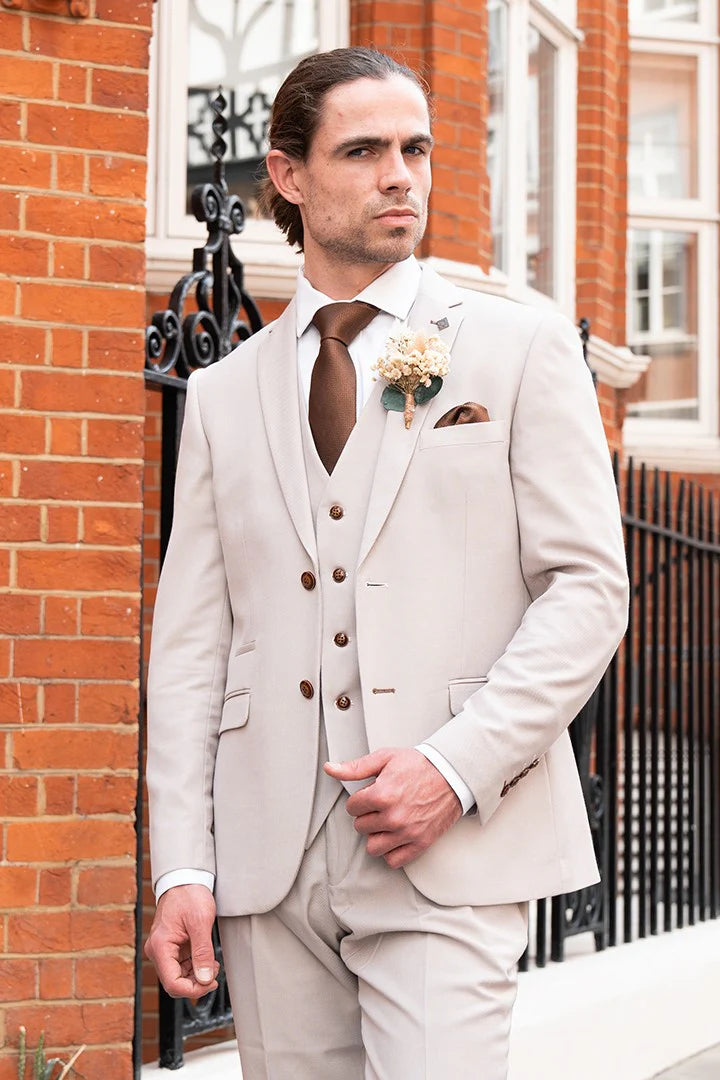 Lifestyle view of a man wearing a stone textured three piece suit with a white shirt, brown tie, matching pocket square and boutonniere beside a brick building.