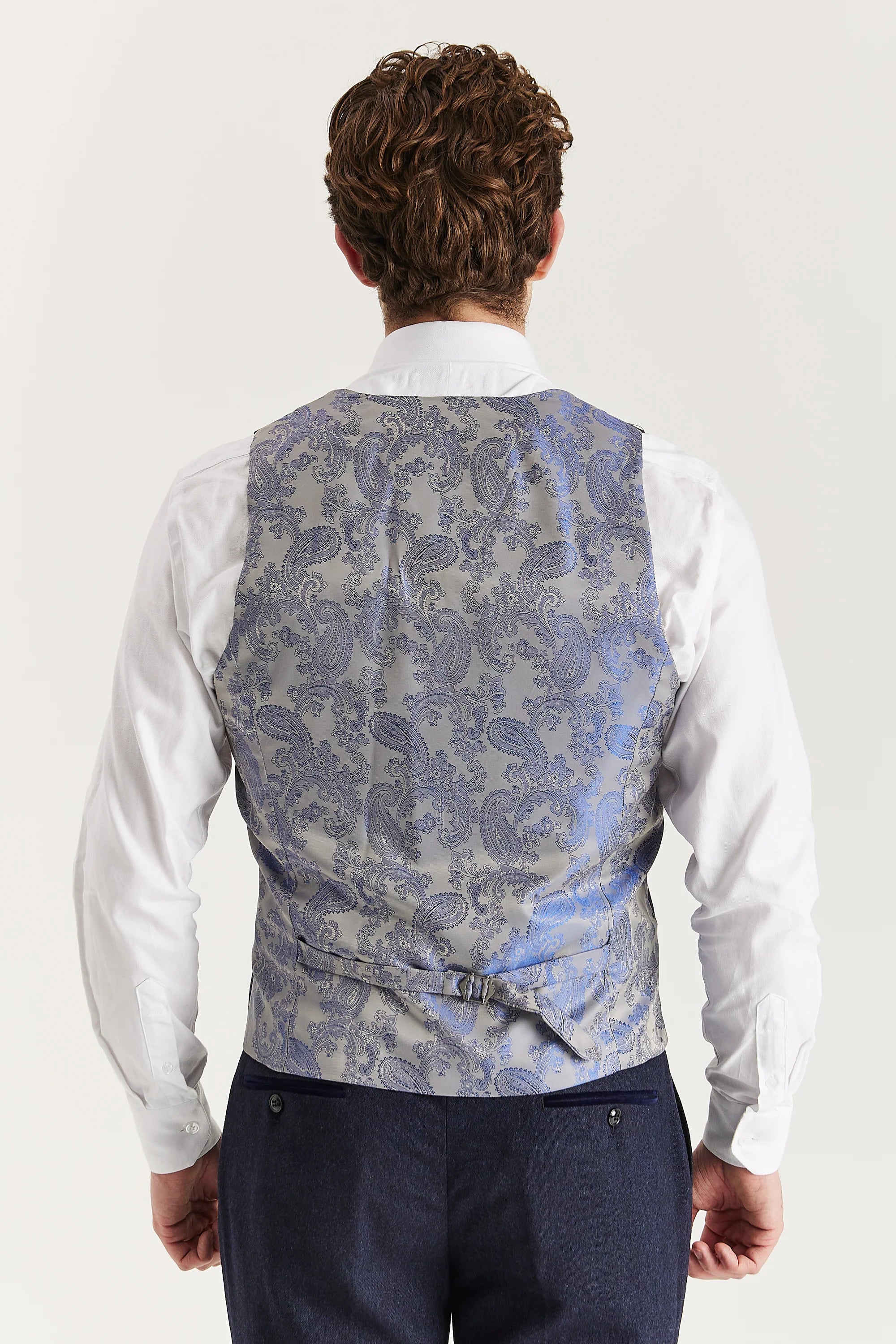 Rear view of the navy waistcoat with a silver paisley back panel and adjustable strap, worn with a white shirt and matching trousers.