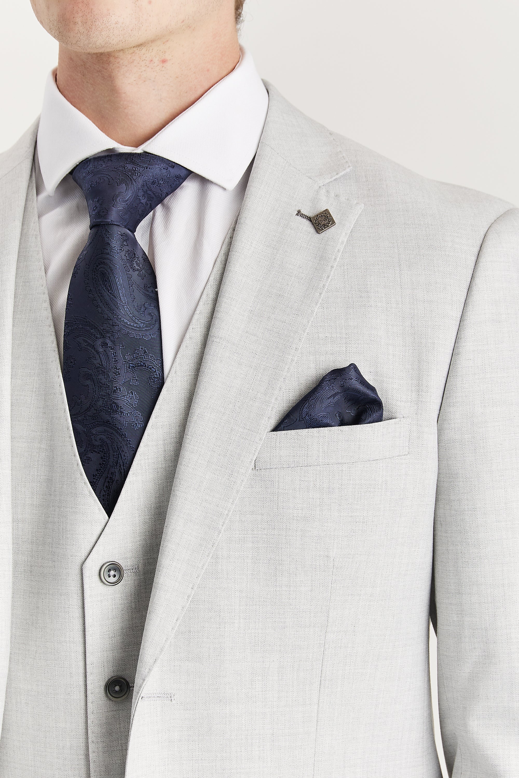 Close-up view of the silver blazer showing the chest pocket, pick stitching, navy paisley tie, matching pocket square and visible lapel pin.