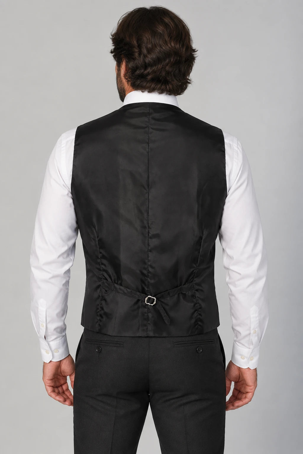 Rear view of a man wearing a black satin waistcoat over a white dress shirt and black trousers, set against a solid gray studio background.
